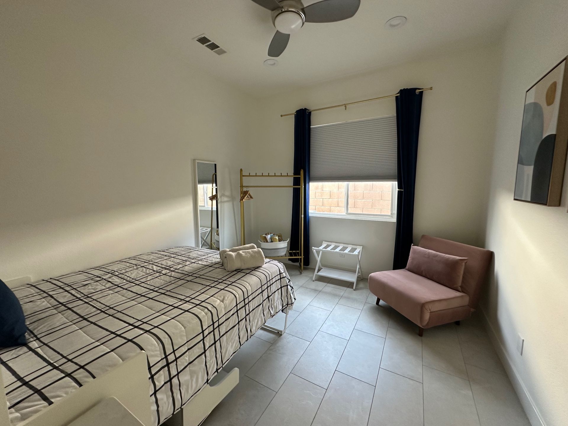A bedroom with a bed, small couch, window, and coat rack.  White walls and flooring.