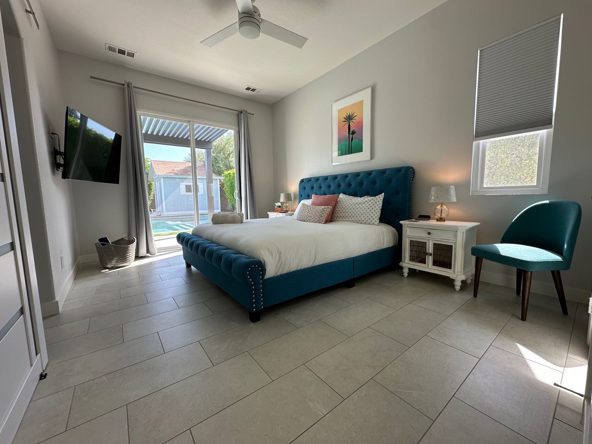 Bedroom with a blue tufted bed, white bedside table, teal chair, and sliding glass door to a pool.