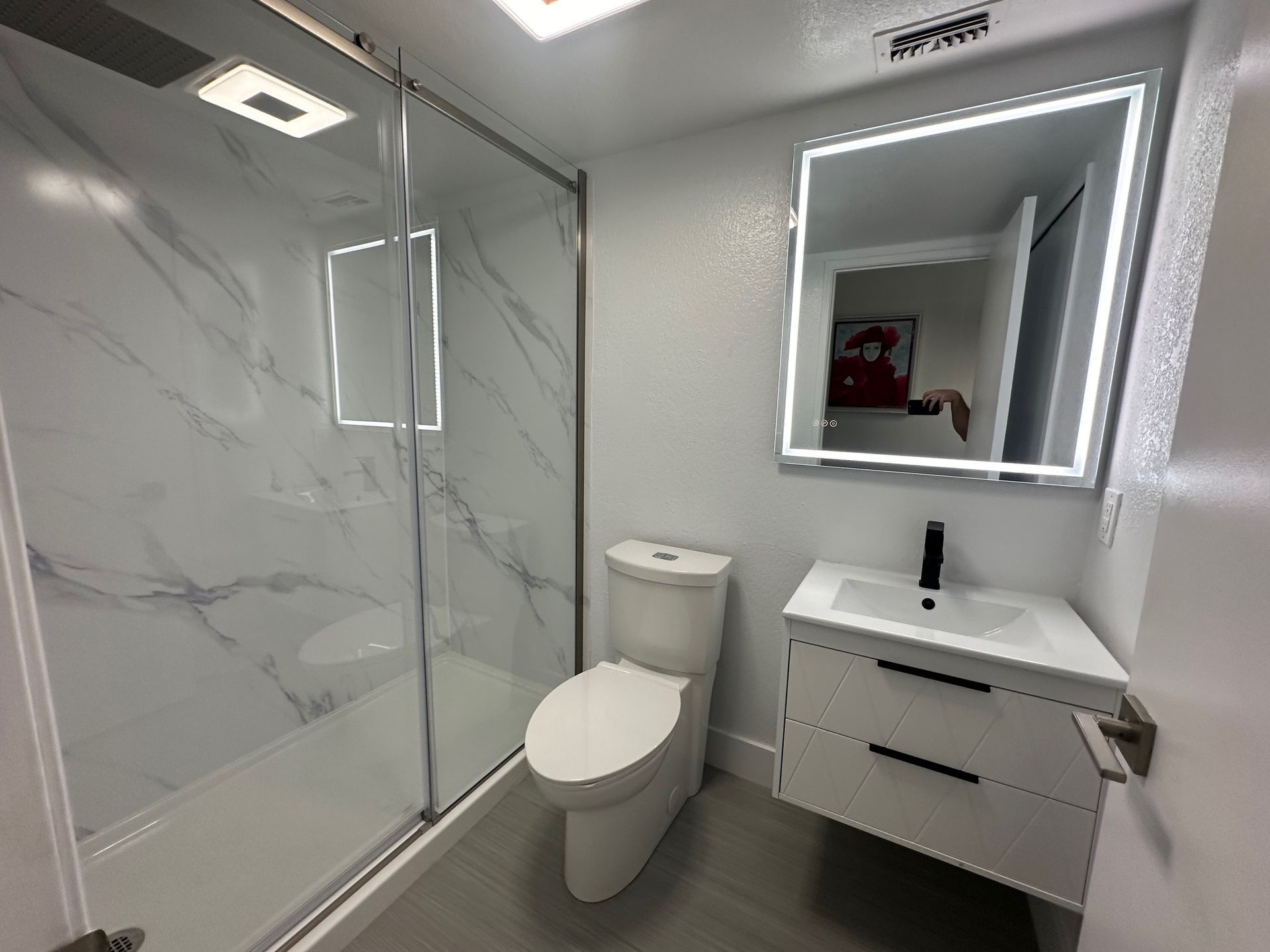 Modern bathroom with white and marble tile shower, floating vanity, toilet, and lighted mirror.