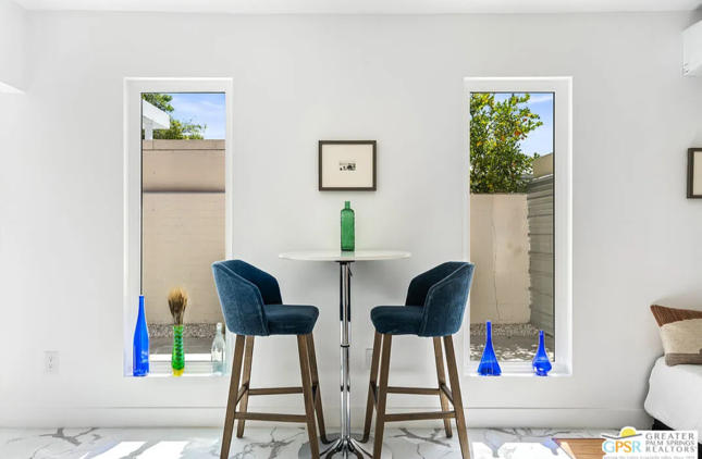 Small white dining area with two blue velvet bar stools and a round table between two windows