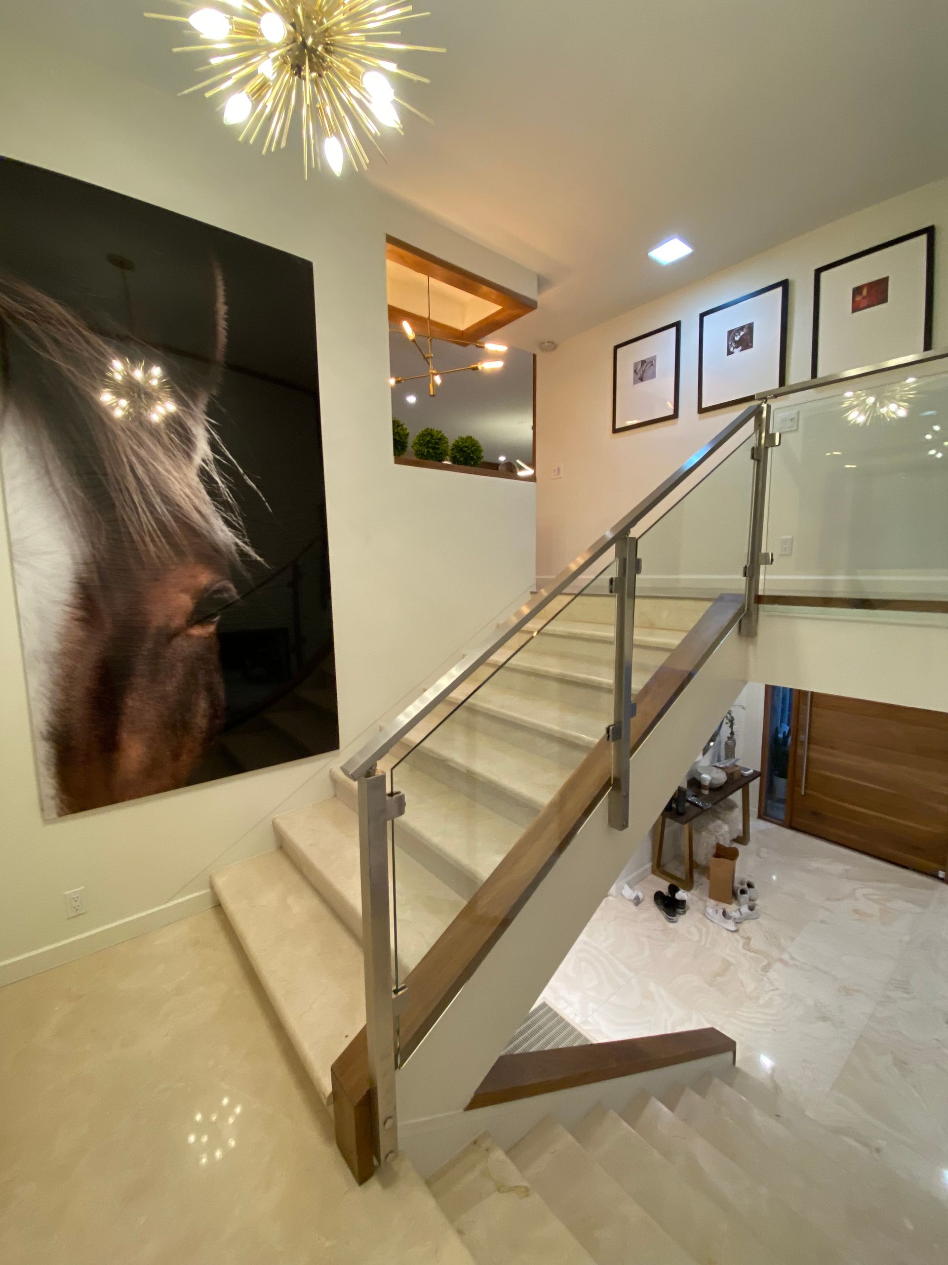 Staircase with glass railing, horse artwork, framed prints, and chandelier in a well-lit interior.