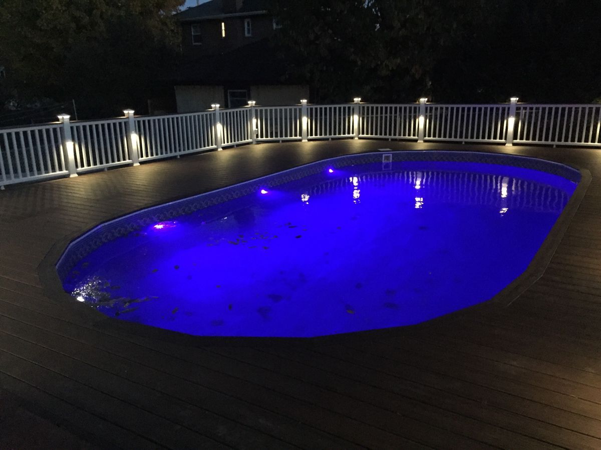 Night view of an illuminated oval pool surrounded by a deck and a white railing with lights.