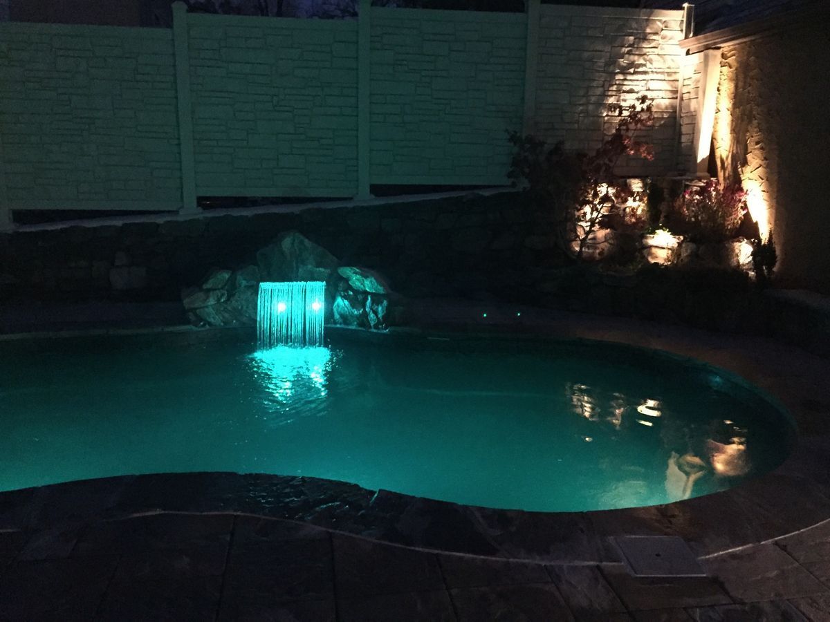 Nighttime pool with illuminated turquoise water and waterfall feature.