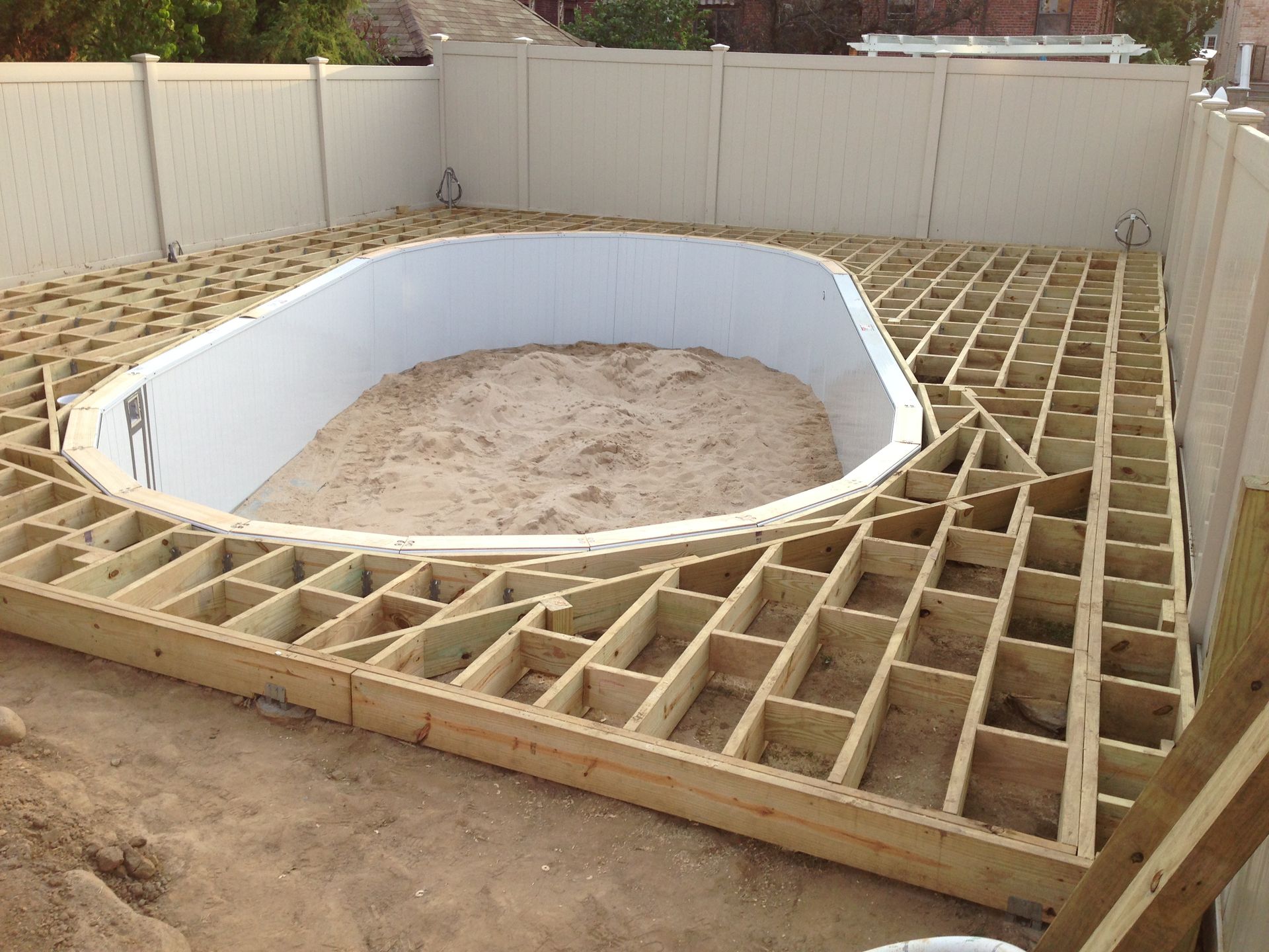 Wooden deck frame around an oval above-ground pool. White vinyl fence surrounds the yard.