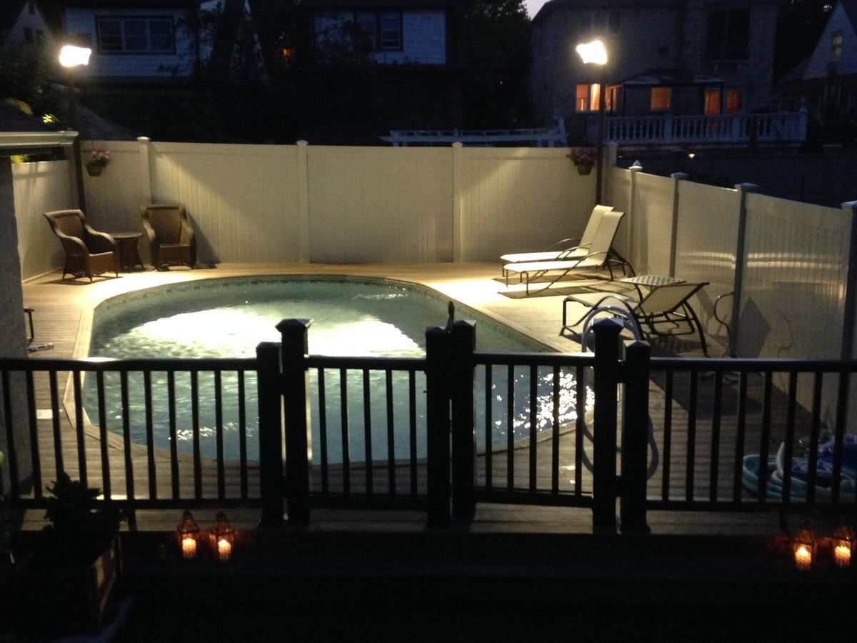 Night view of a swimming pool surrounded by a white fence and lit by spotlights. Chairs are visible nearby.
