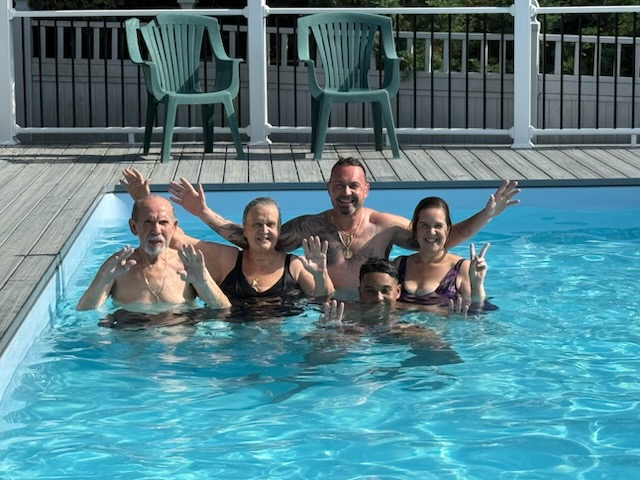 Group of people in a pool, smiling and waving. Wooden deck and green chairs in the background. Sunny day.