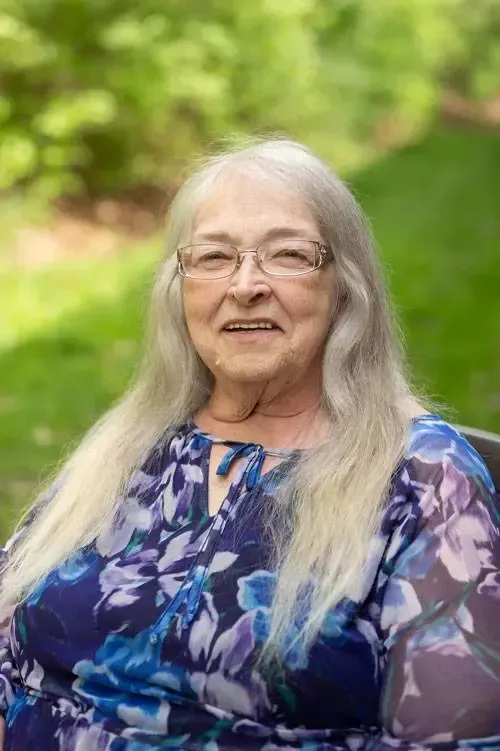 An elderly woman wearing glasses and a blue floral dress is sitting in a wheelchair.