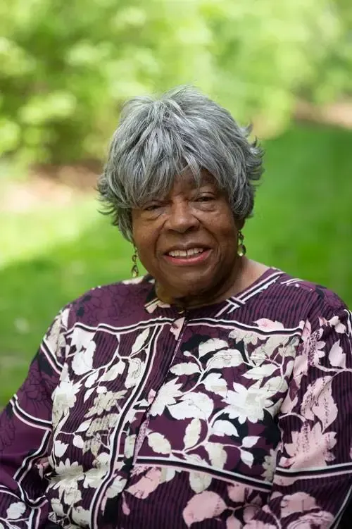 A woman with gray hair is wearing a purple floral shirt and smiling.