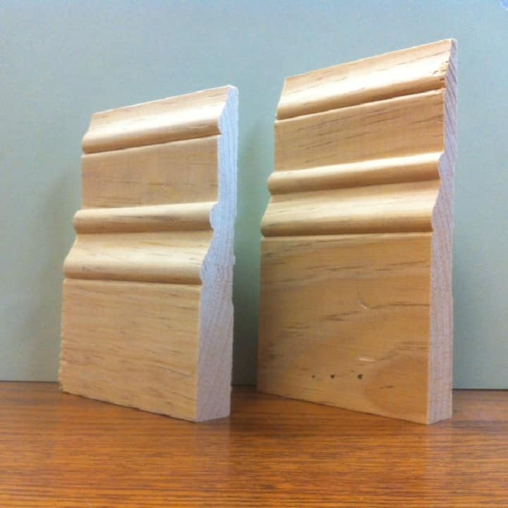 Two wooden baseboard samples, light brown color, with curved and straight edges, resting on a brown surface.