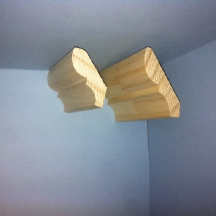 Two pieces of unfinished wooden crown molding at a corner, against blue and white walls.
