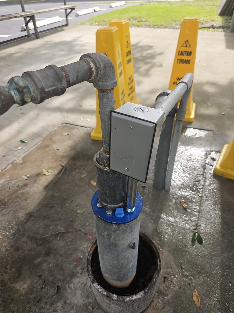 A wellhead with metal piping and electrical box, surrounded by caution signs on concrete.