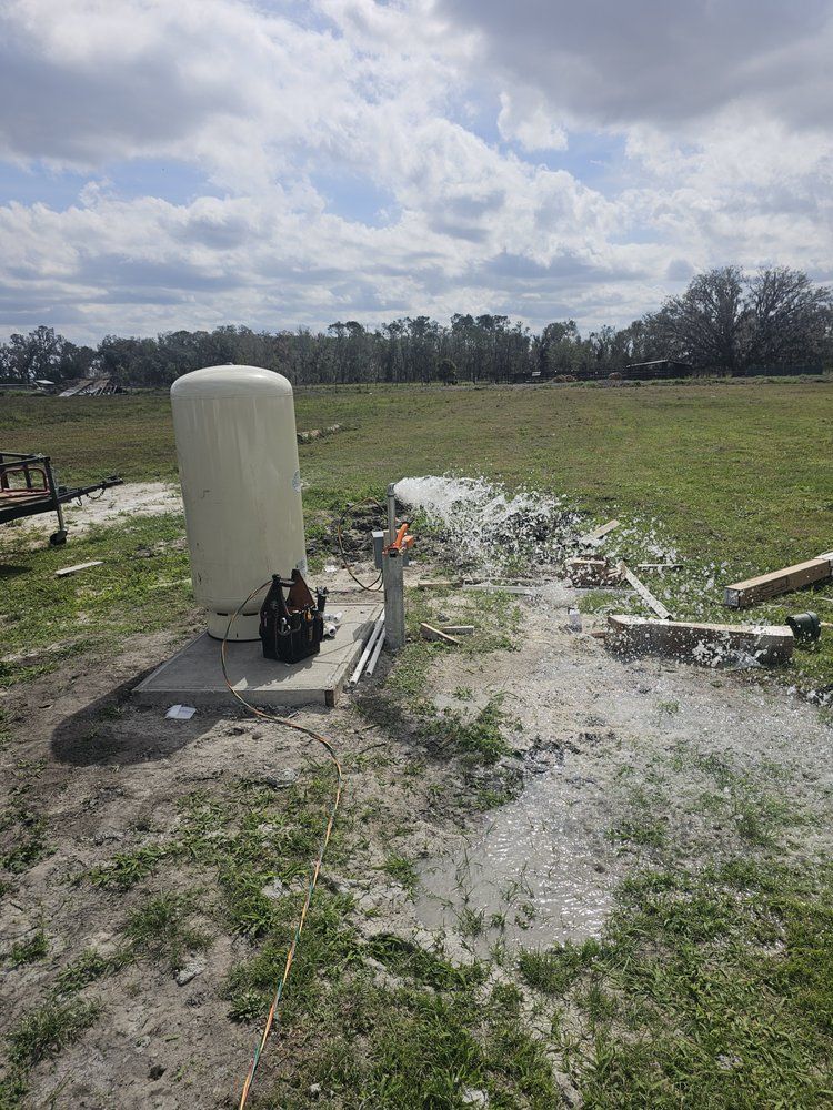 Well pump spraying water onto the ground in a field. Beige tank and equipment on a concrete pad.