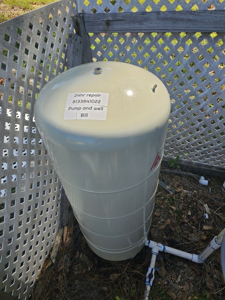 Tan water pressure tank outside, near a white lattice fence, with connecting pipes.