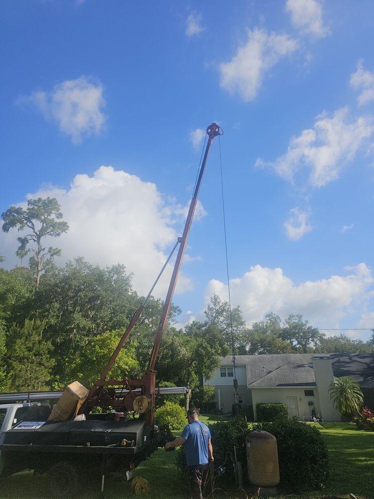 Red drilling rig against a blue sky, operated by a person next to a truck. Trees and houses in the background.