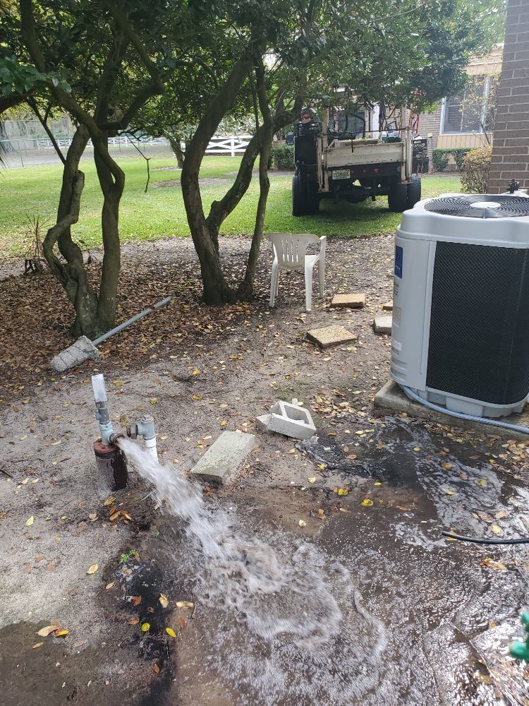 Water gushes from a well on a muddy ground next to an air conditioner unit. A truck is in the background.