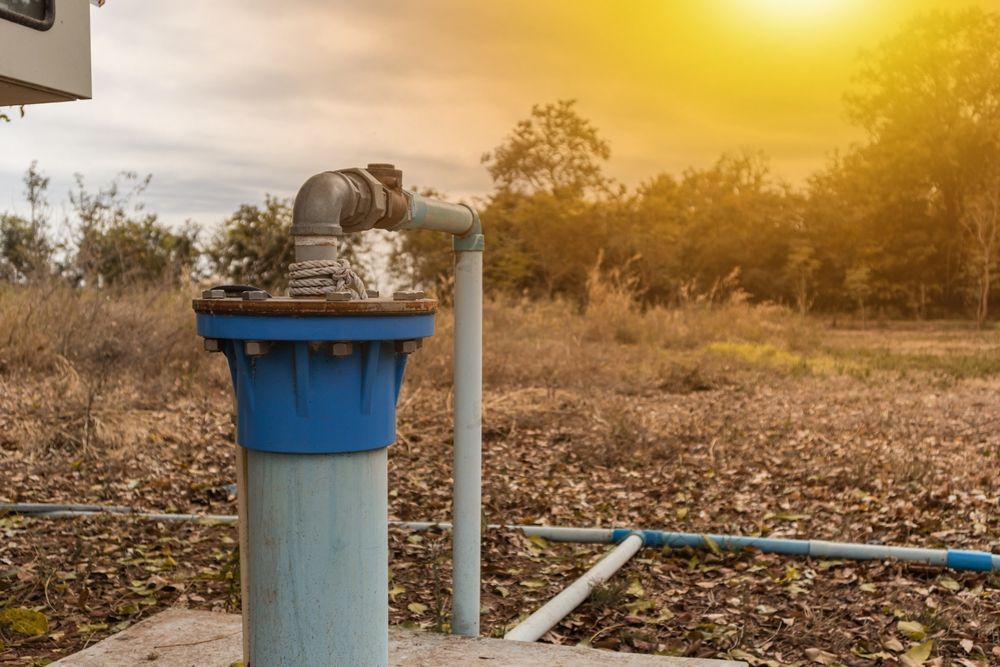 A blue well casing with a pipe, spraying water, in a field with dry vegetation and a cloudy sky.