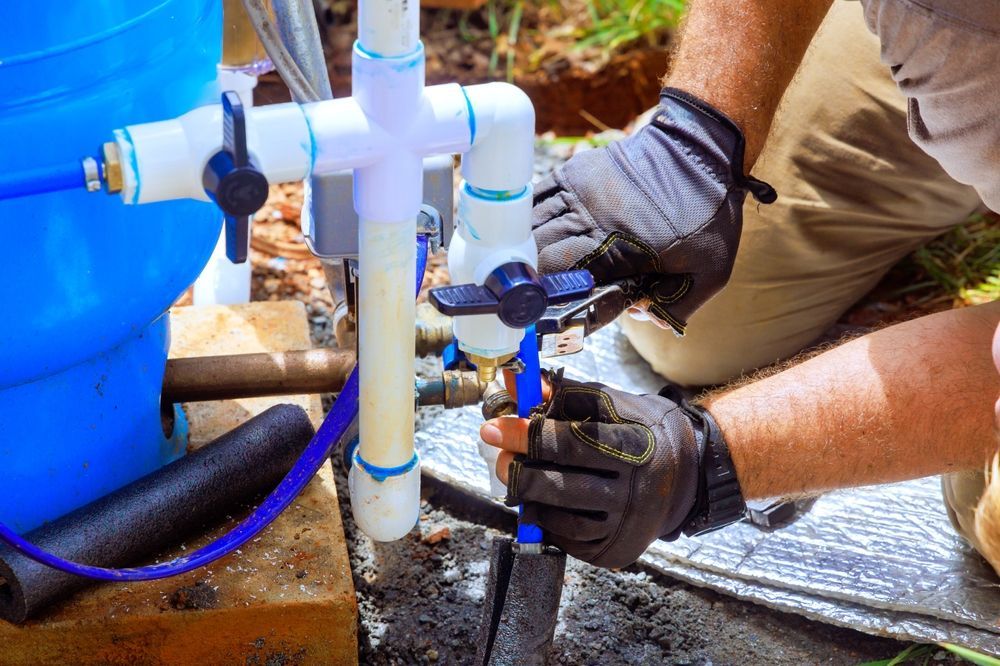 Person in gloves working on plumbing near a blue tank and pipes.