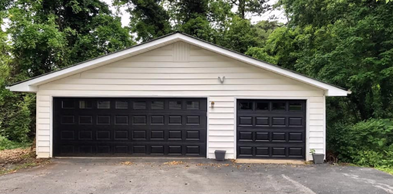 A white garage with black garage doors is surrounded by trees.