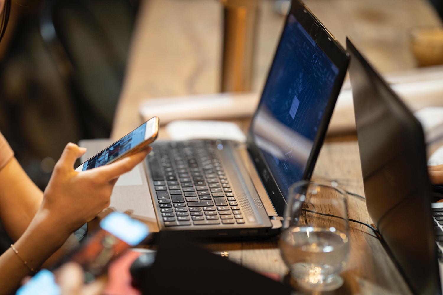 A woman is sitting at a table using a laptop and a cell phone.