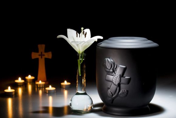 Memorial candle setup with a white lily, wooden cross, and black urn on a dark table