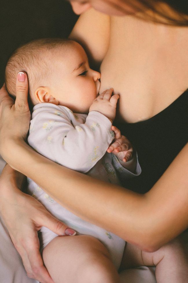 Baby breastfeeding, held by parent. Parent wearing black top, baby in onesie. Dark background.