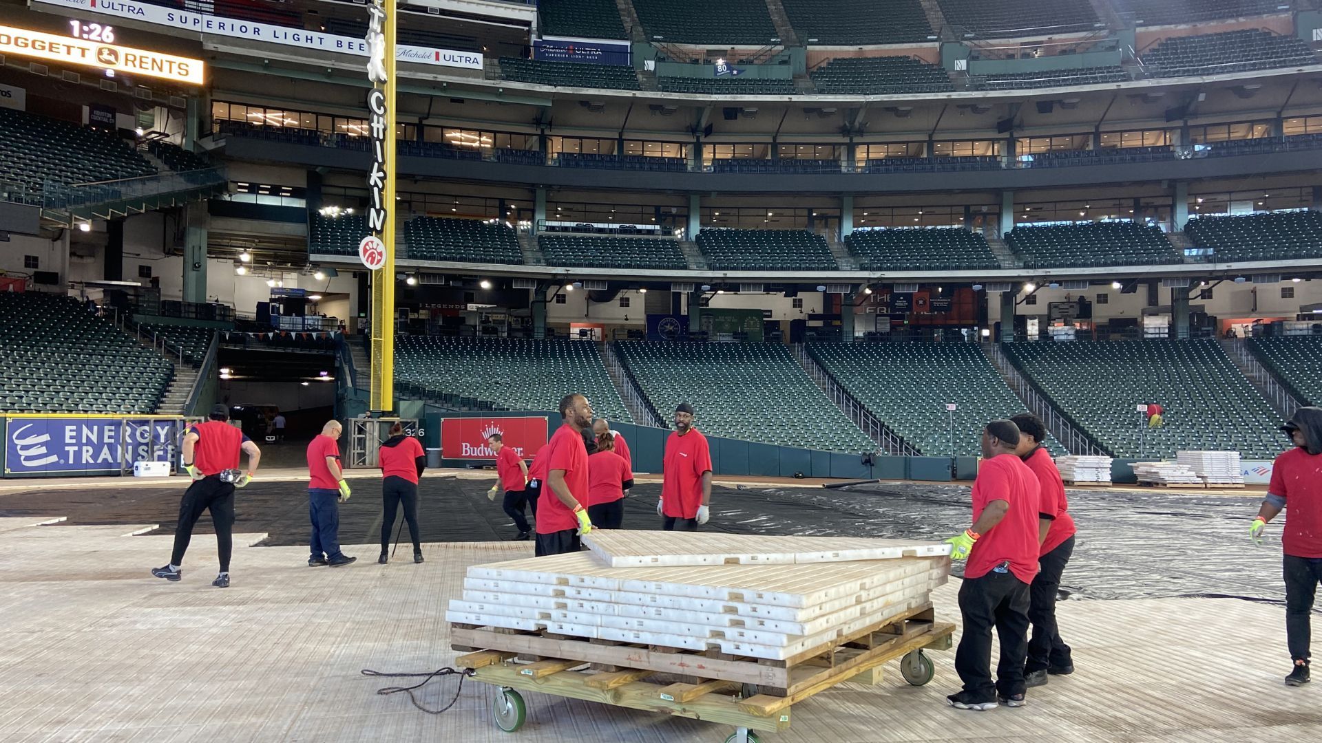 Workers in red jackets move equipment across a baseball stadium field under the stands.