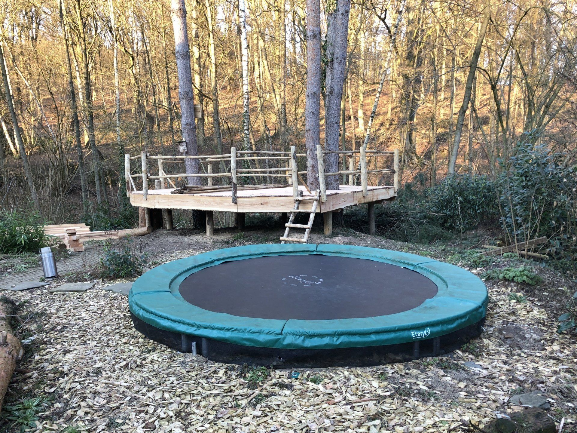 Midden in het bos staat een trampoline naast een houten terras.
