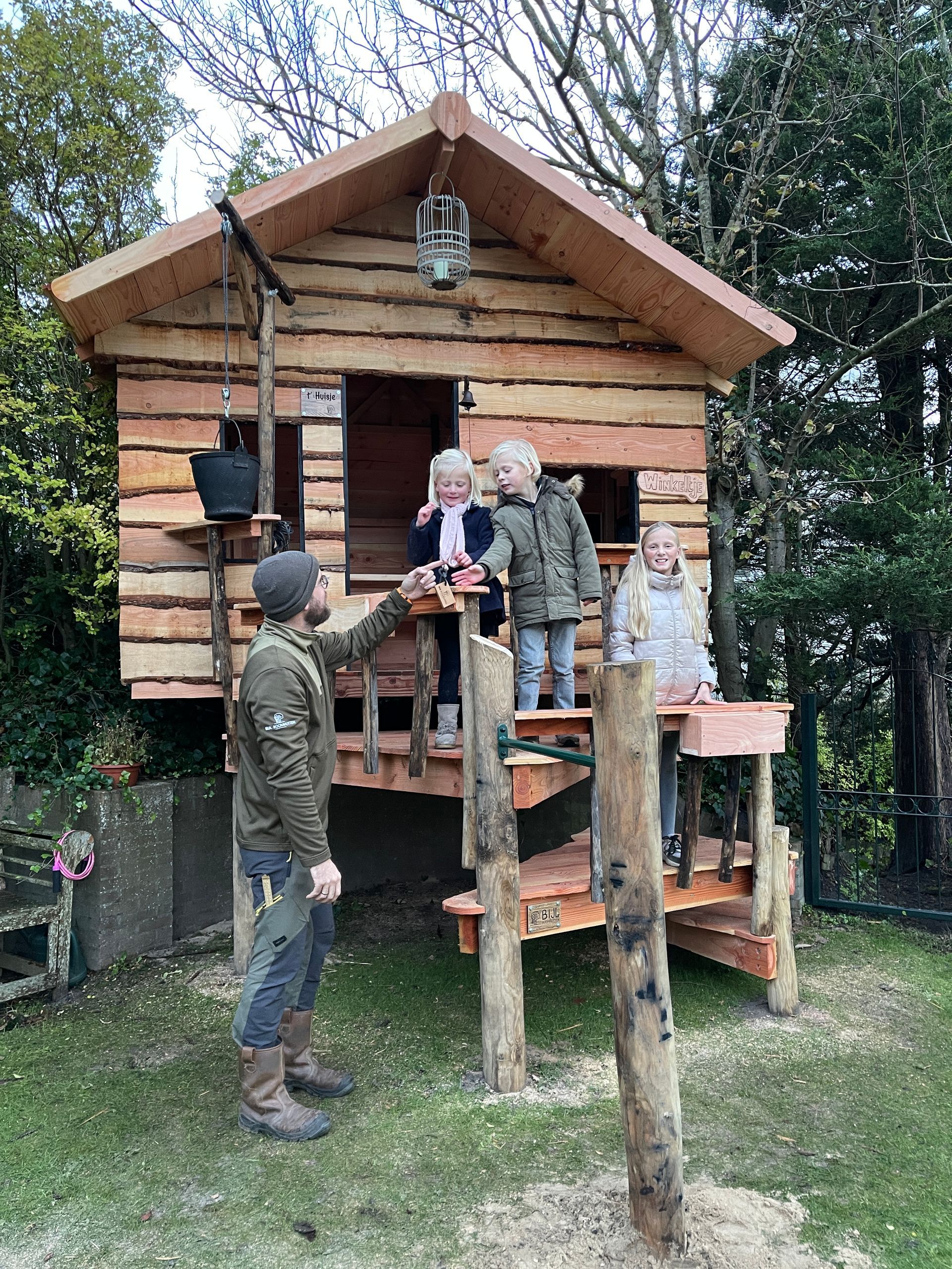 Een man en twee kinderen staan ​​voor een houten boomhut.