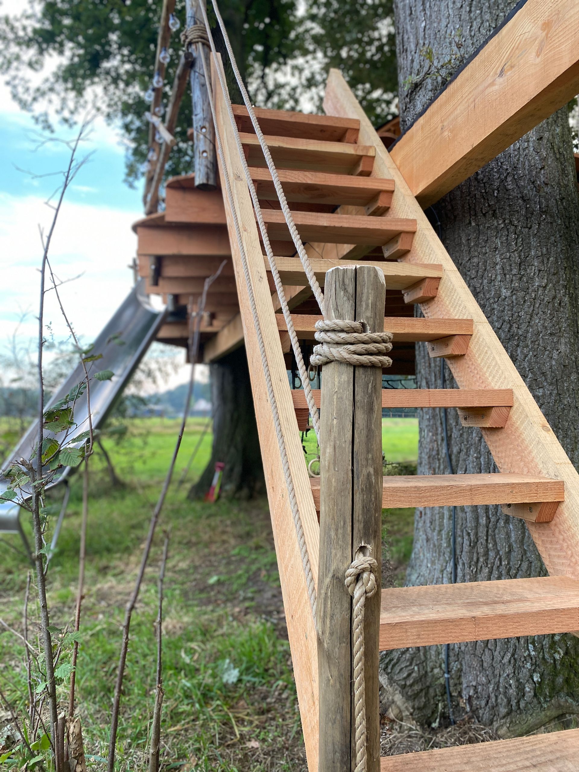 Een houten trap die leidt naar een boomhut met een glijbaan op de achtergrond.
