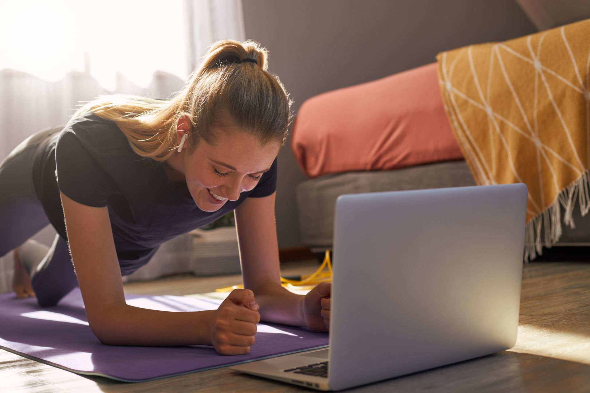 A woman is doing a plank on a yoga mat while looking at a laptop.