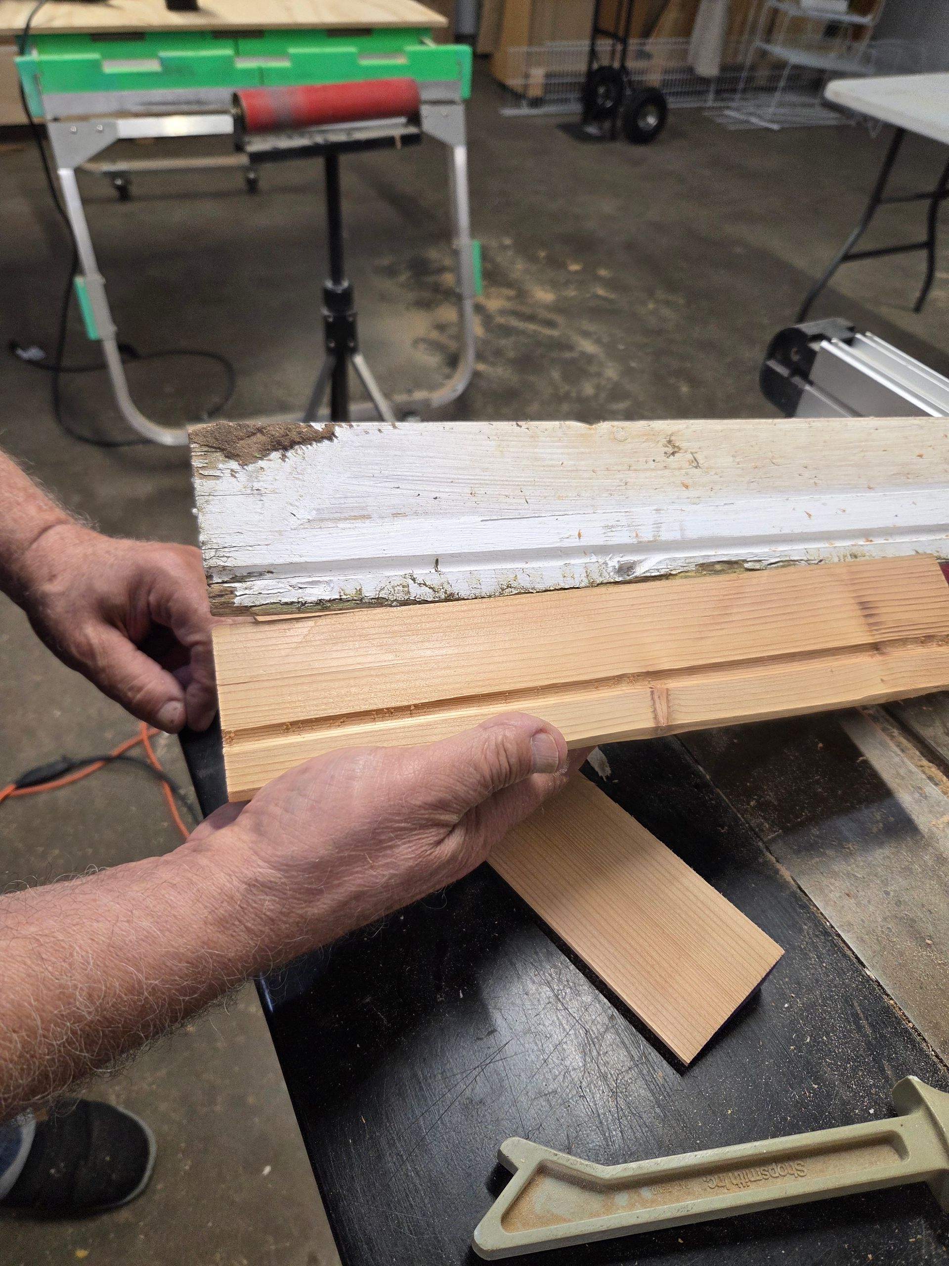 A person's hands holding a piece of raw wood against an existing painted trim board in a workshop.