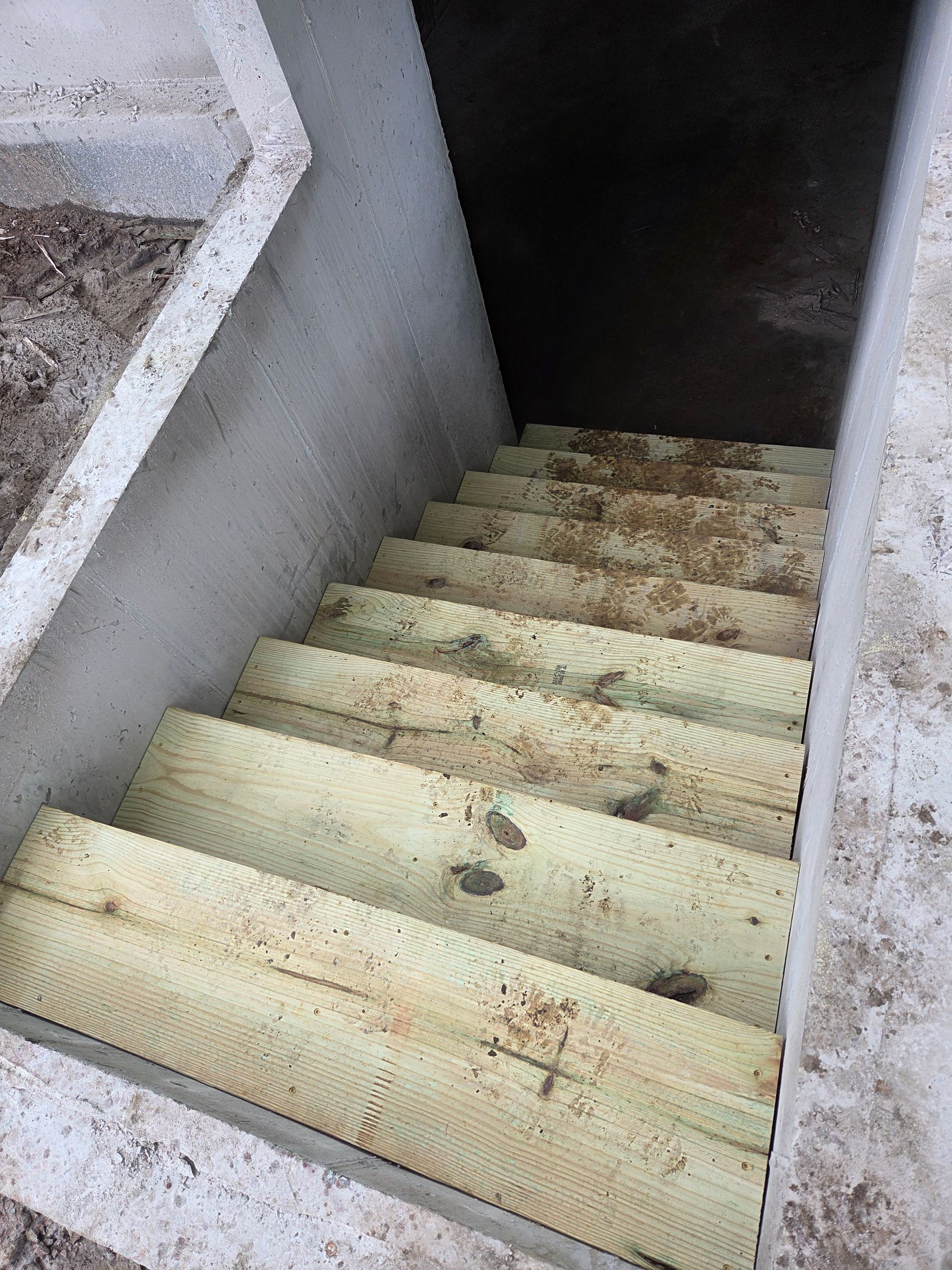 A set of light-colored wooden stairs leading down into a dark basement entrance between two concrete walls.