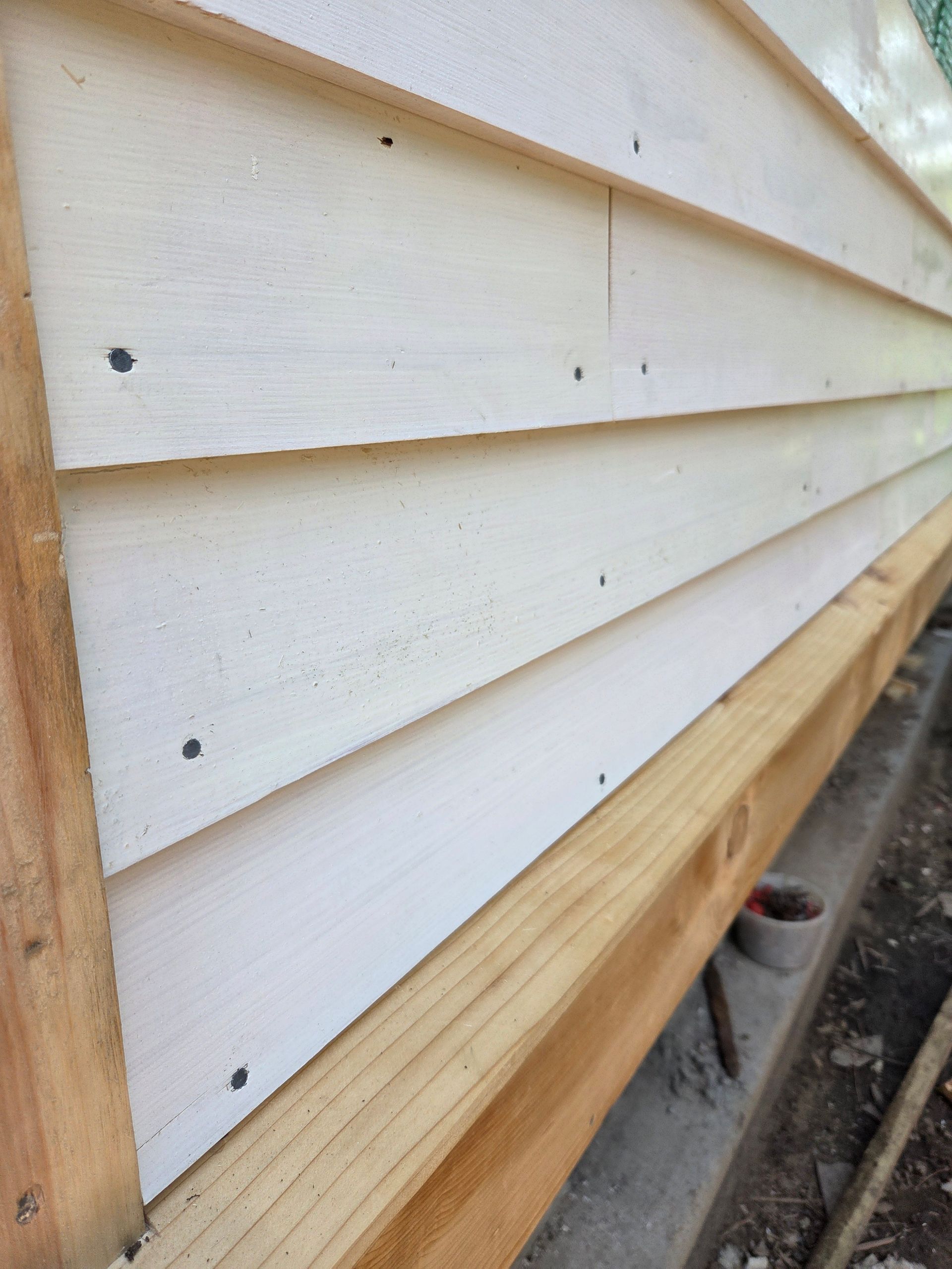 A close-up view of white lap siding installed on a wooden frame, showing nail heads and the unfinished base beam.