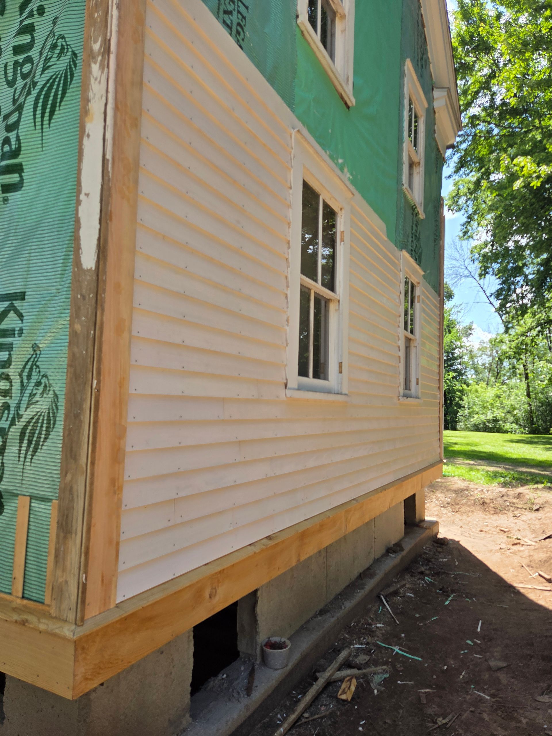 A side view of a house undergoing renovations, showing light beige siding being installed over green, insulated sheathing.