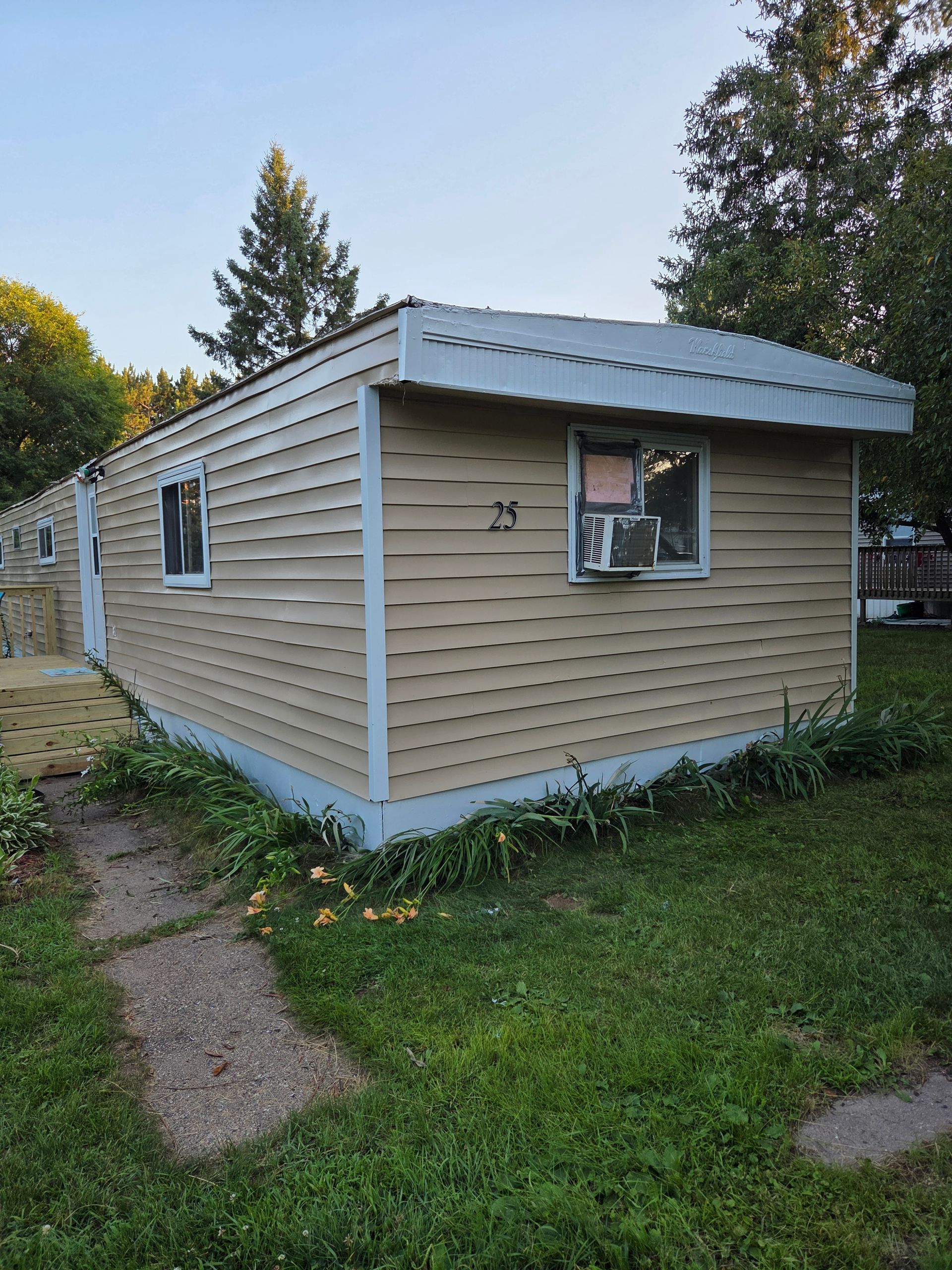 A beige mobile home with a light blue foundation and an air conditioning unit in the window, set in a grassy yard.