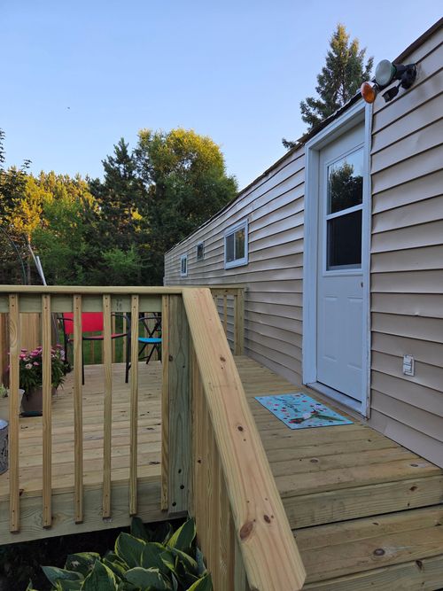 A wooden deck with a railing leads to the white door of a light-colored mobile home, set against a backdrop of trees.