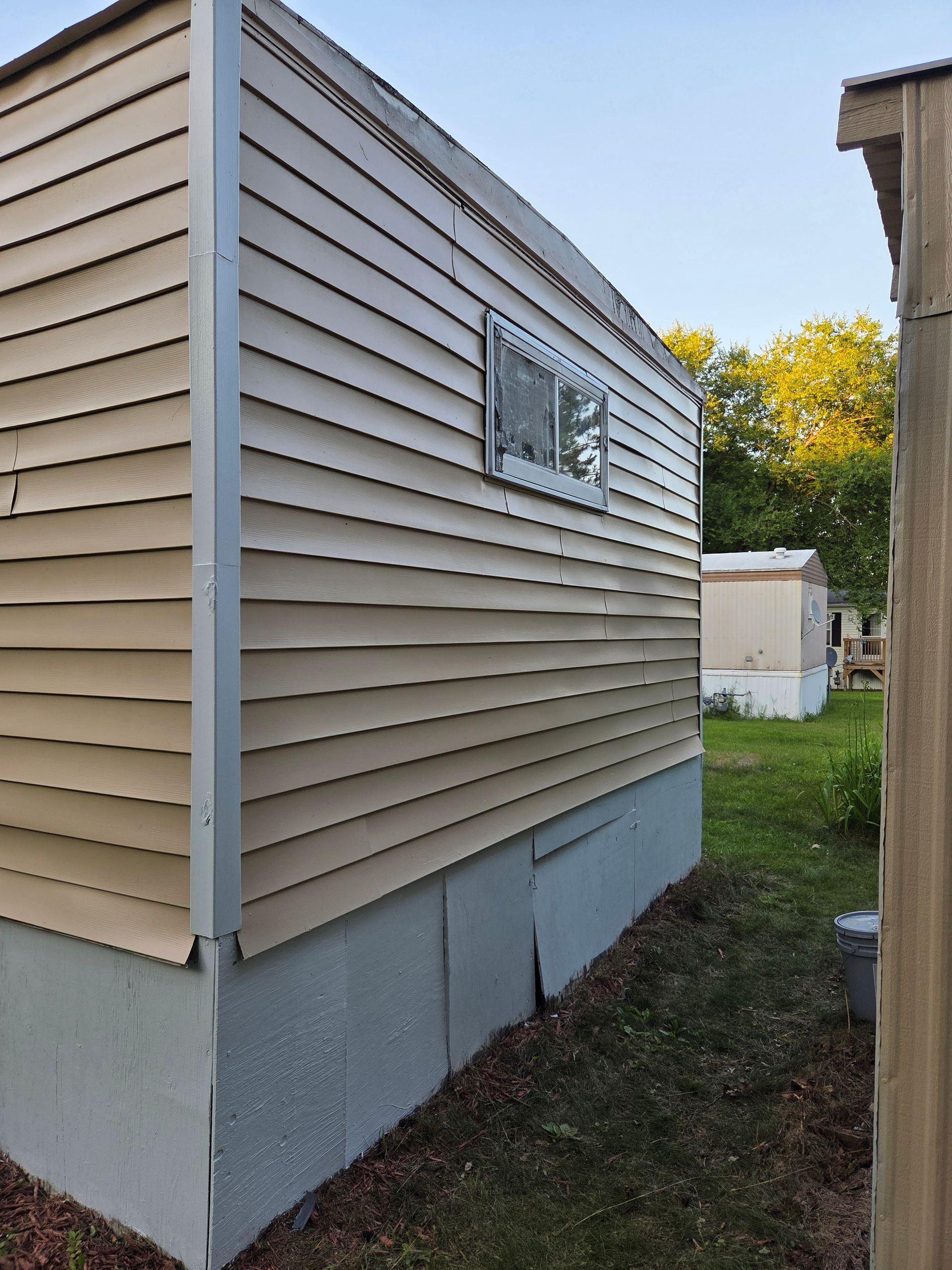 A tan mobile home with a small window and gray skirting, situated in a yard with trees in the background.