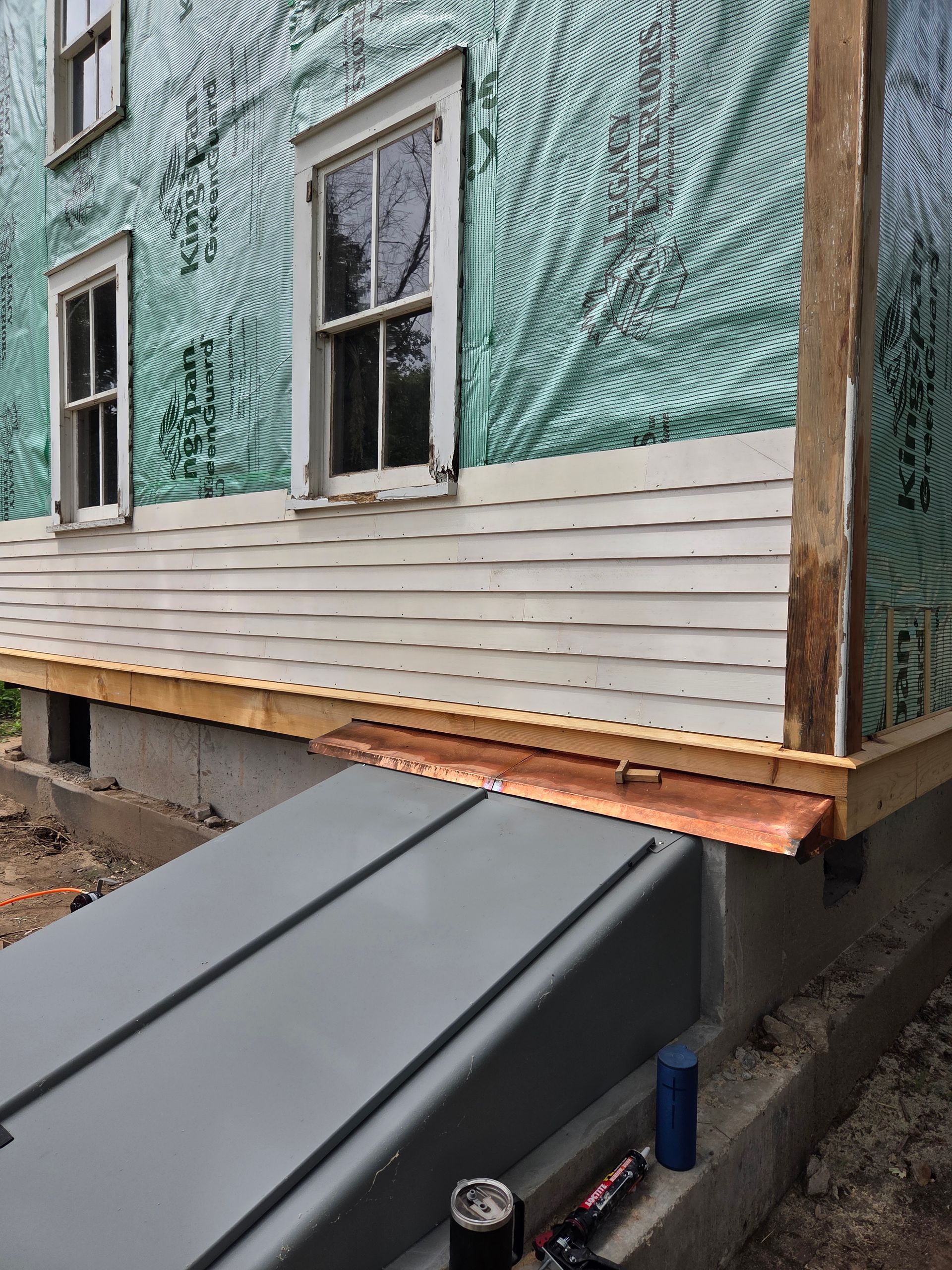 A house exterior under construction with white siding, a teal vapor barrier, and a new copper flashing over a cellar door.