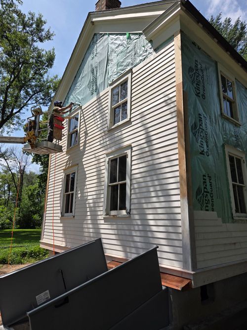 Workers in a lift platform install white siding on a house undergoing exterior renovation.