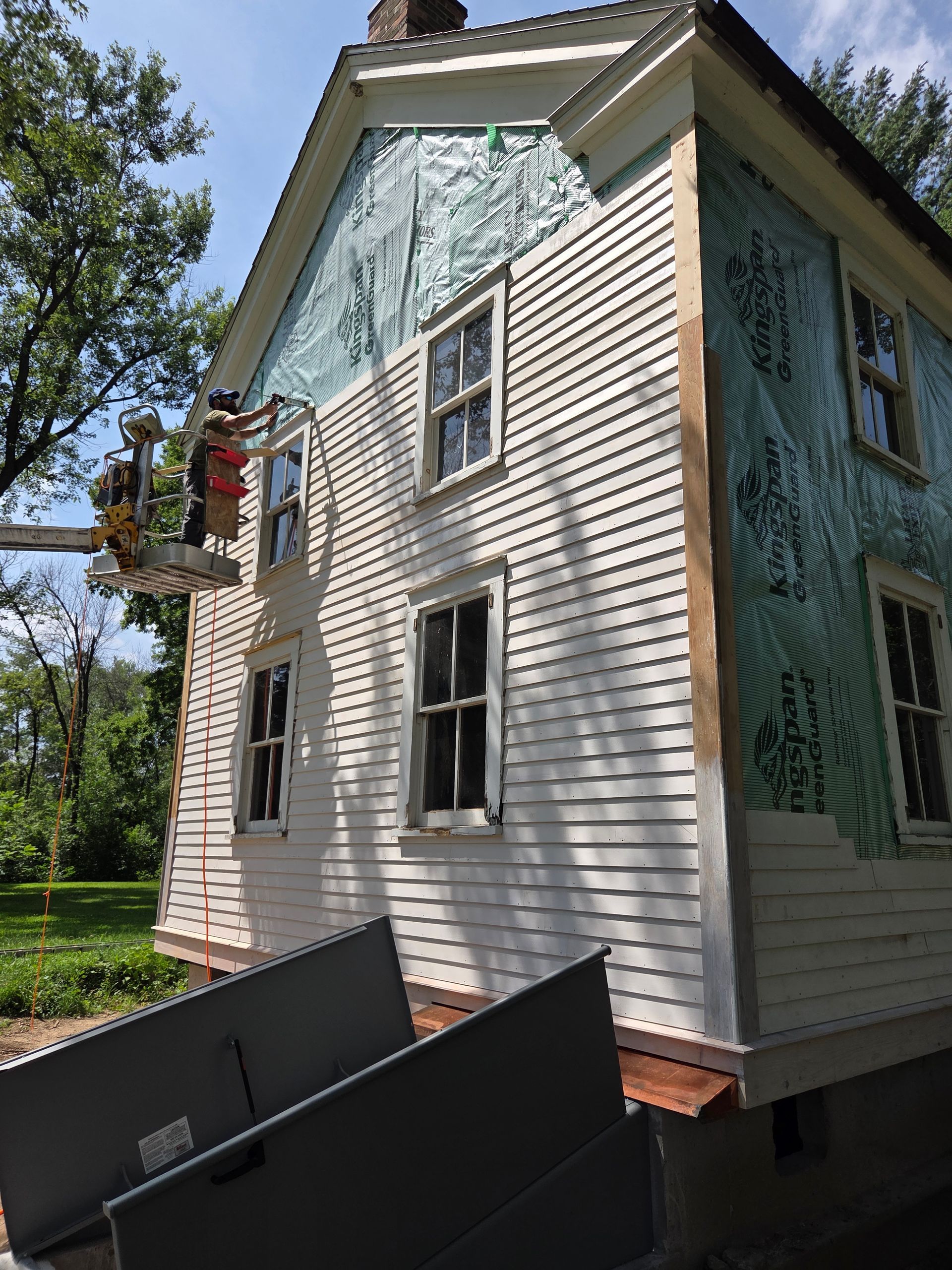 Workers in a lift platform install white siding on a house with visible green protective underlayment.