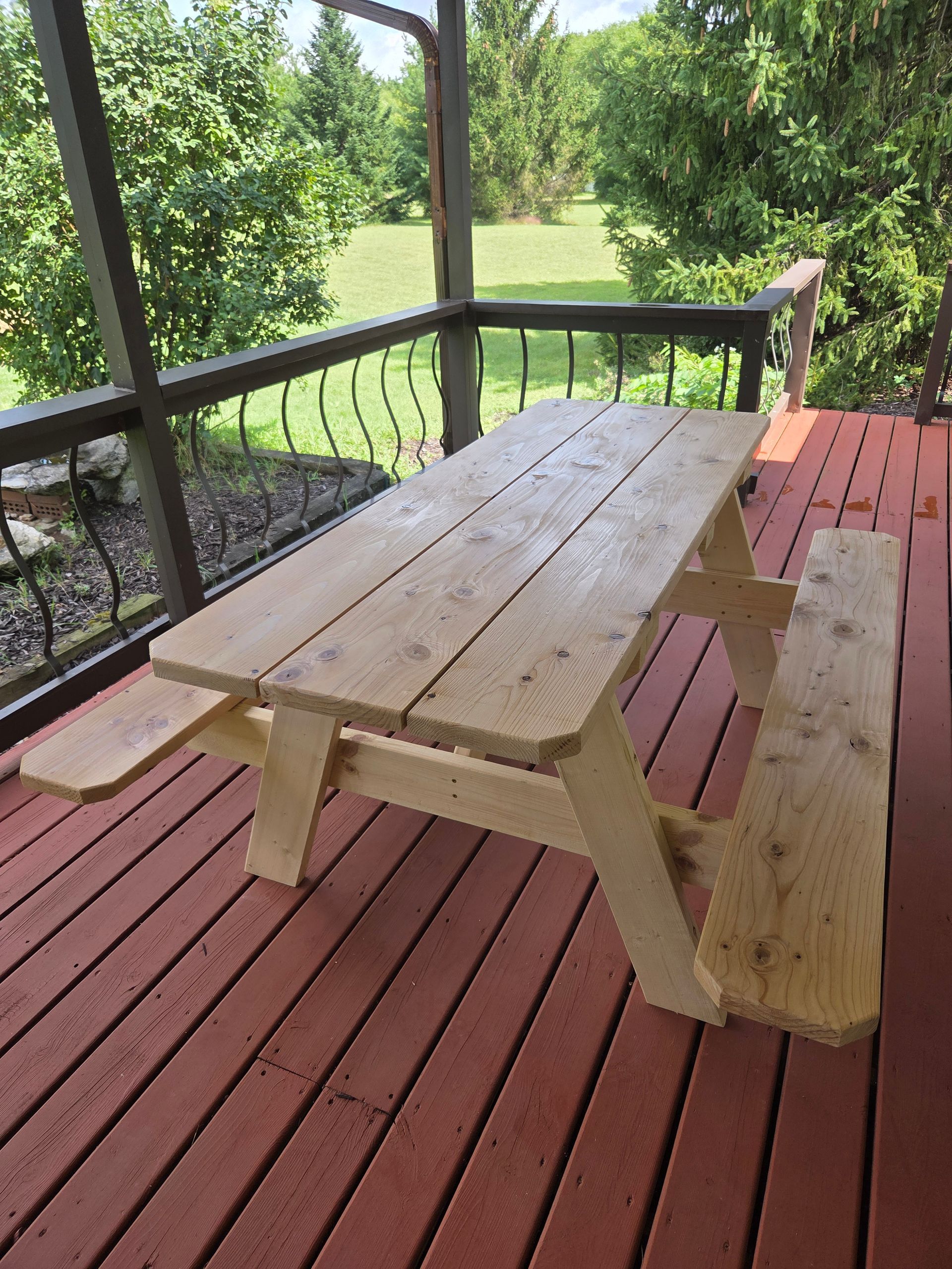 A wooden picnic table sits on a red-painted outdoor deck overlooking a grassy yard with trees.