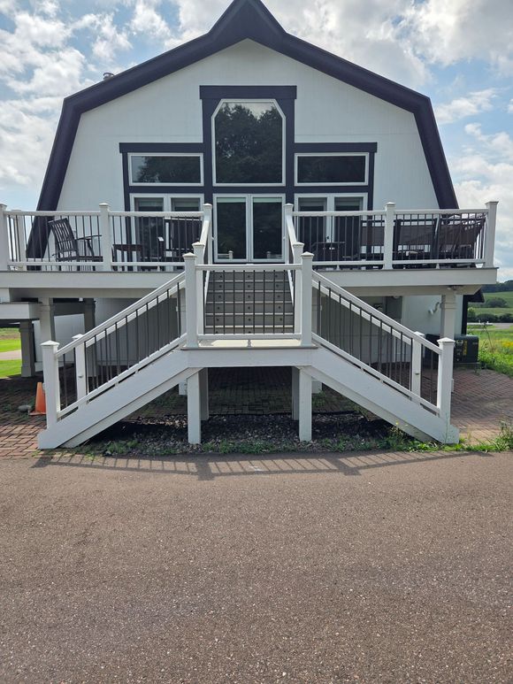A white, barn-style house with dark trim, featuring a large deck accessed by a split staircase over a gravel driveway.