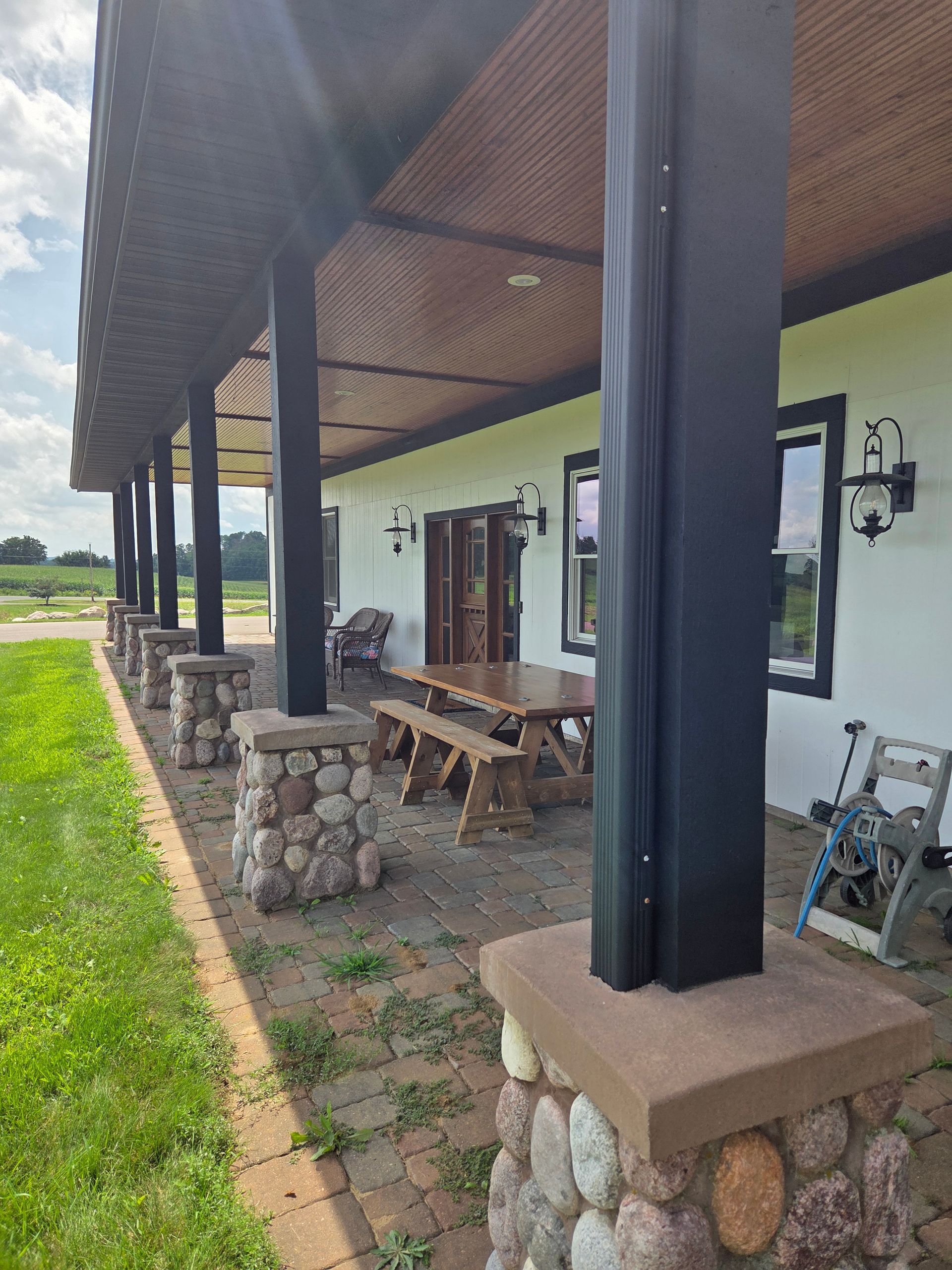 A covered stone patio with dark support pillars, a wooden picnic table, and exterior wall lanterns along a white building.