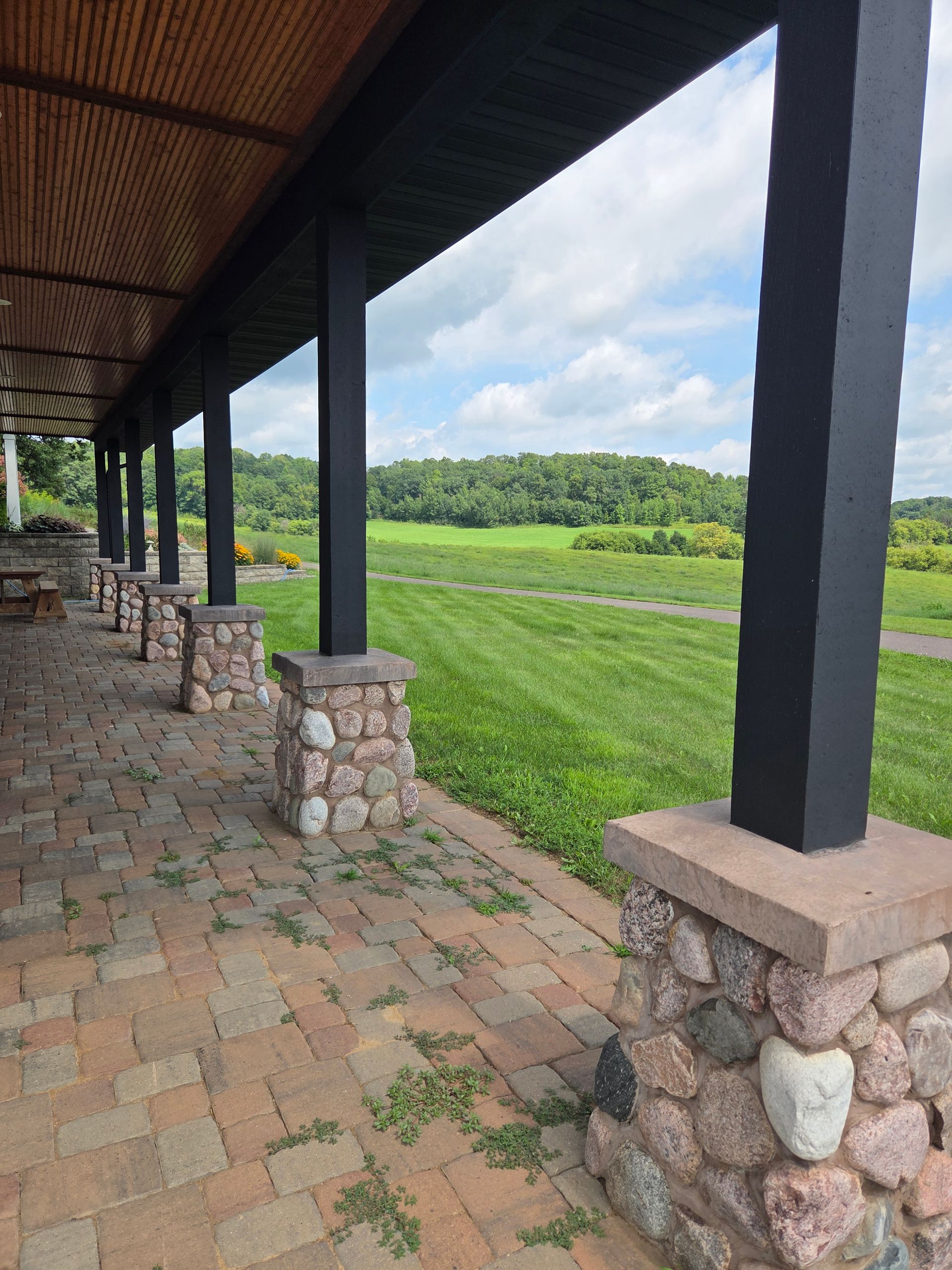 A stone patio with a series of black pillars supporting a roof, overlooking a grassy meadow under a cloudy sky.
