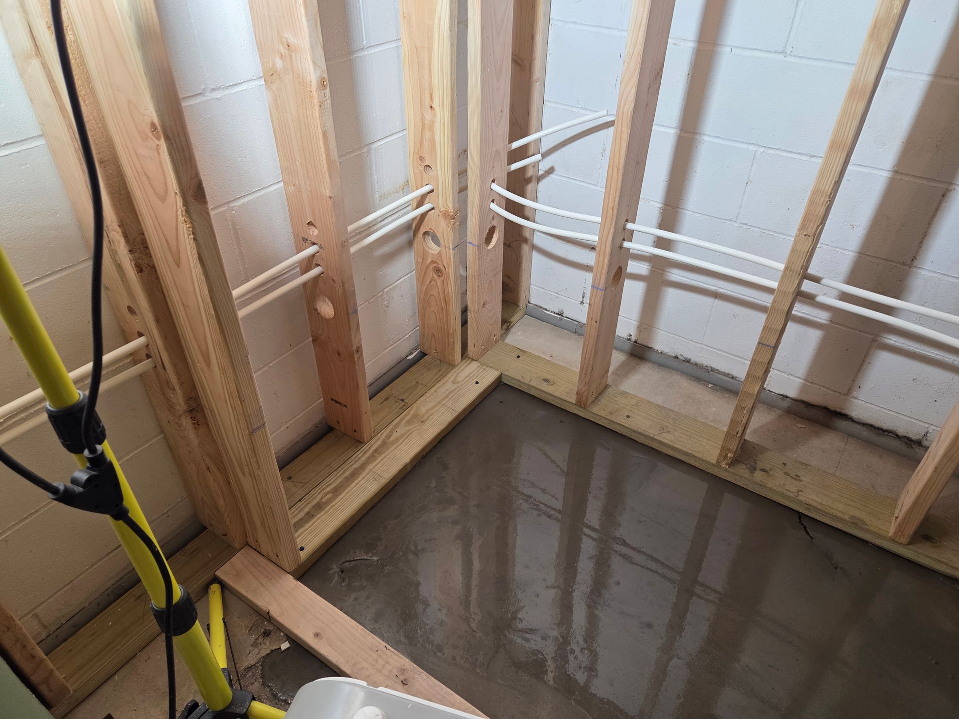 Wooden wall studs installed on a concrete floor with PEX plumbing pipes running through them in a basement corner.