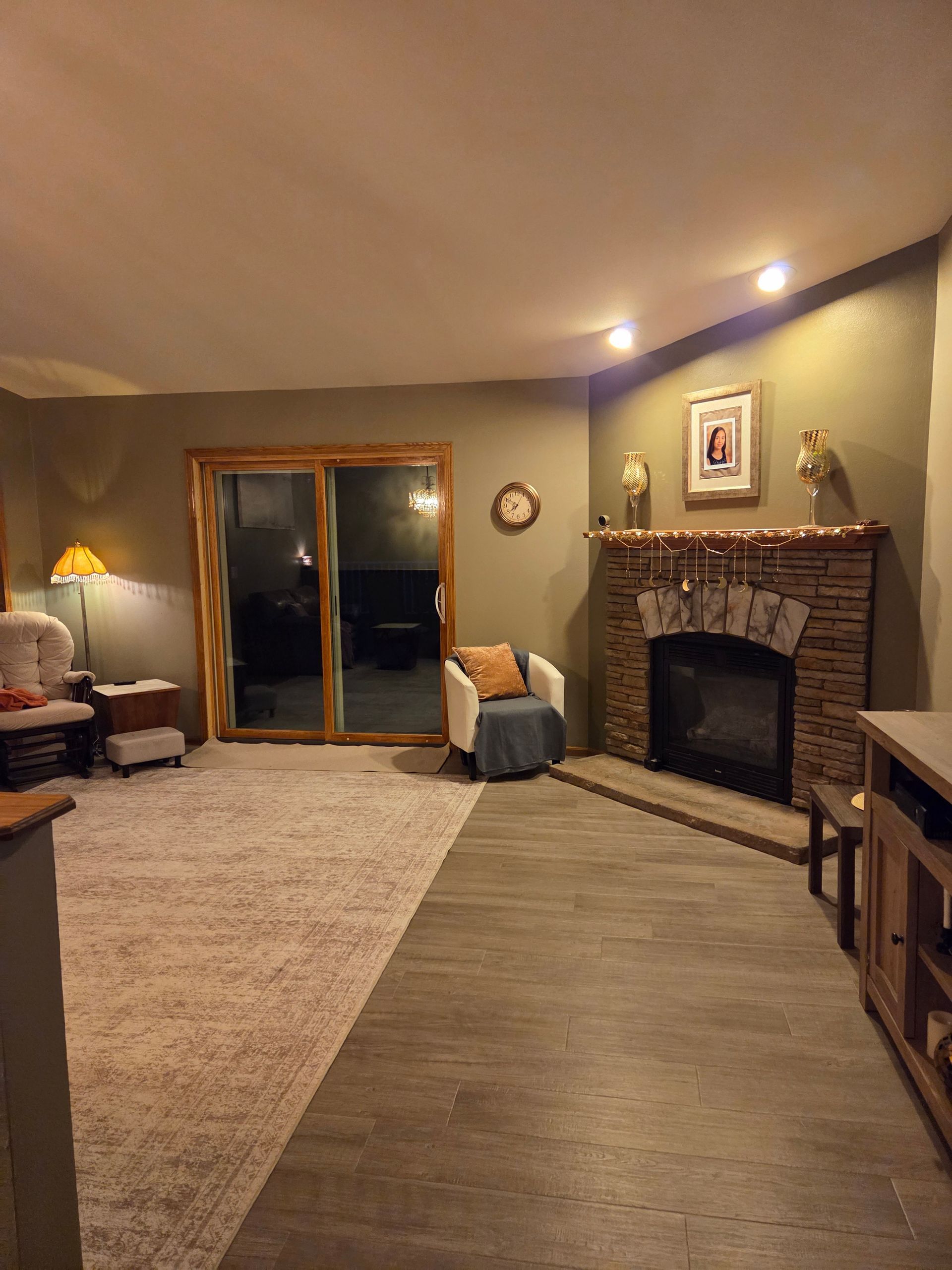 A living room with a stone fireplace, gray wood-look flooring, beige carpet, a sliding door, and two accent chairs.