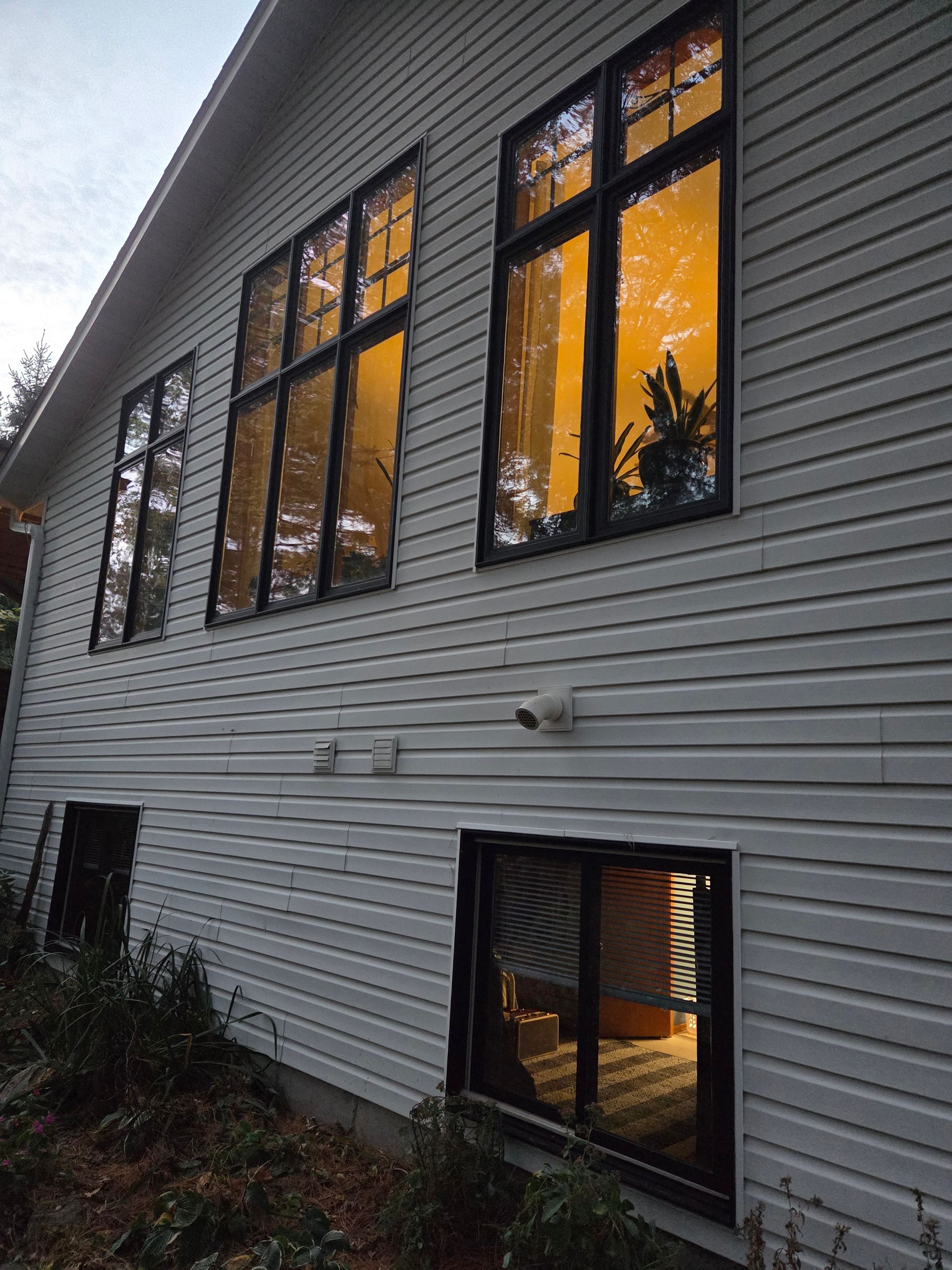 Low-angle view of a white-sided house exterior with tall dark-framed windows reflecting the evening sky and light.