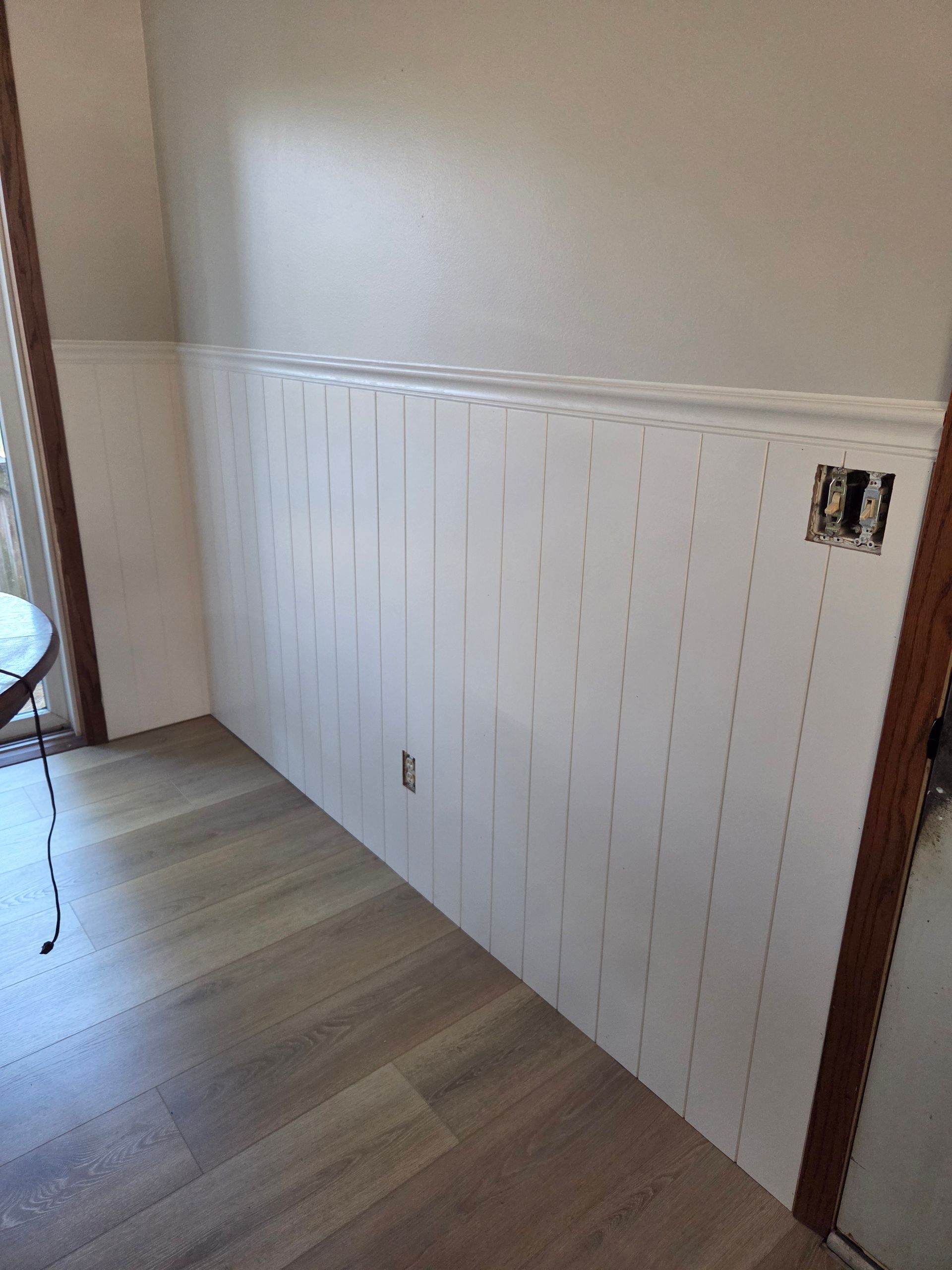 White vertical wainscoting paneling installed on a wall, with a chair rail, light grey wall above, and wood plank flooring.