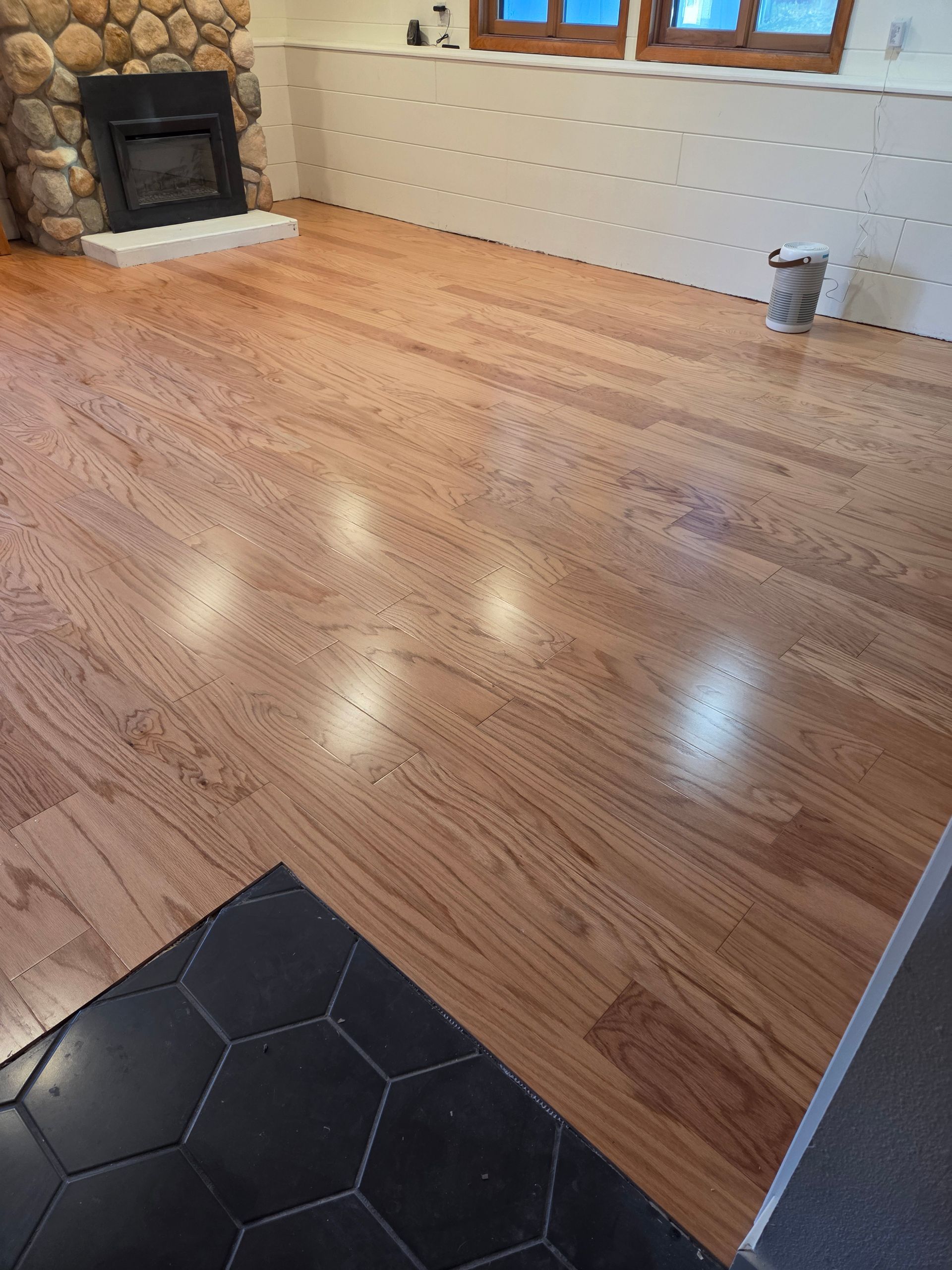 A room with light wood flooring, a stone fireplace, white paneled walls, and a black hexagonal-tiled foreground.