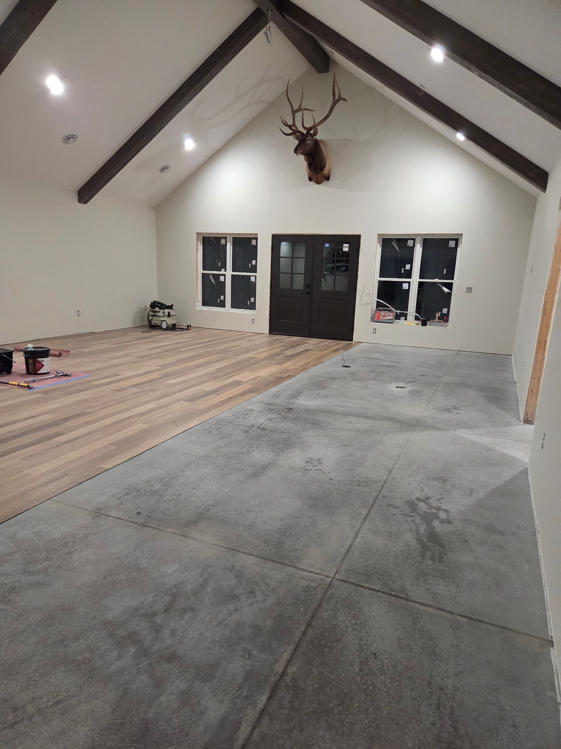 A room with a vaulted ceiling, exposed wooden beams, half-installed wood flooring, and a deer mount above double doors.